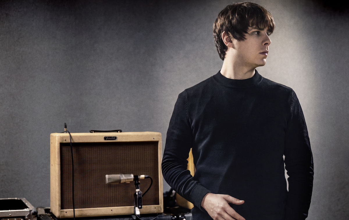 Promotional studio portrait of musician Jake Bugg standing in a black sweater next to a vintage tweed guitar amplifier against a grey backdrop.